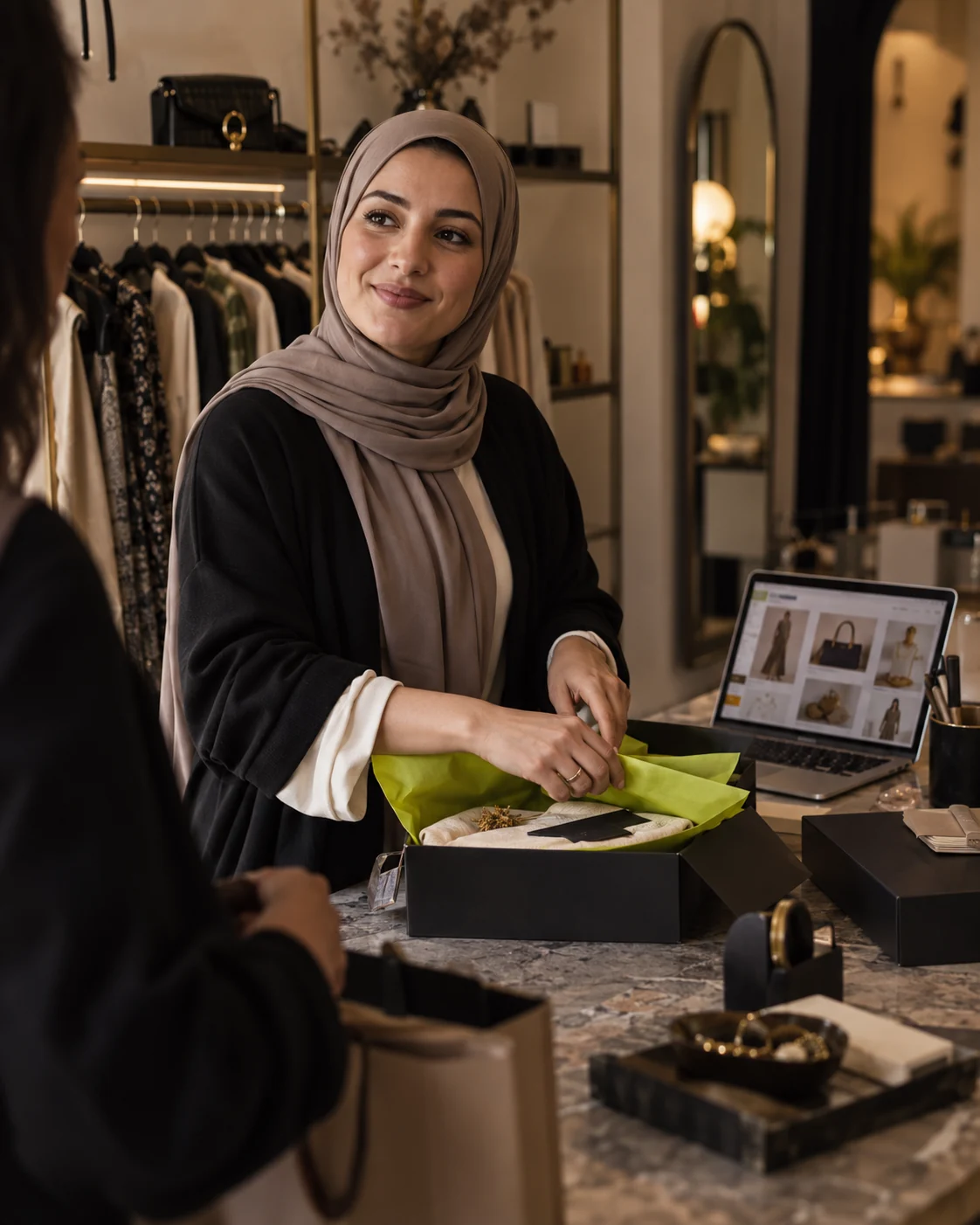 A boutique owner in a taupe hijab packing an order beside her online store on a laptop.